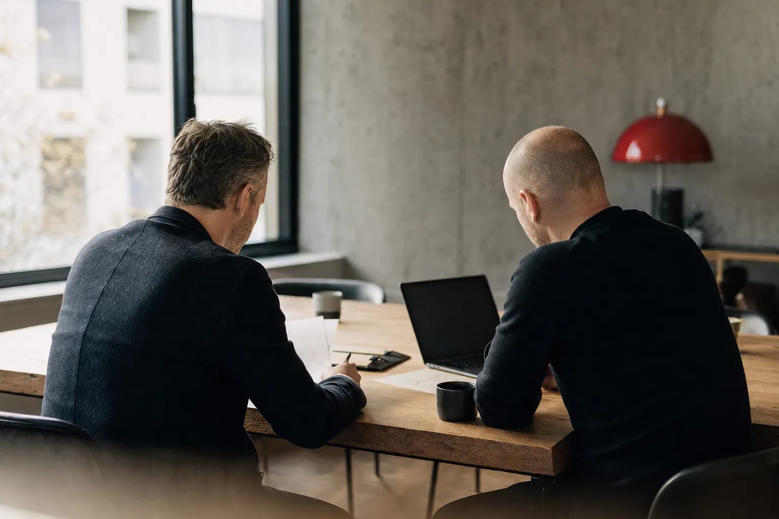 Zwei Personen arbeiten nebeneinander an einem langen Holztisch mit Notizen und Laptop.