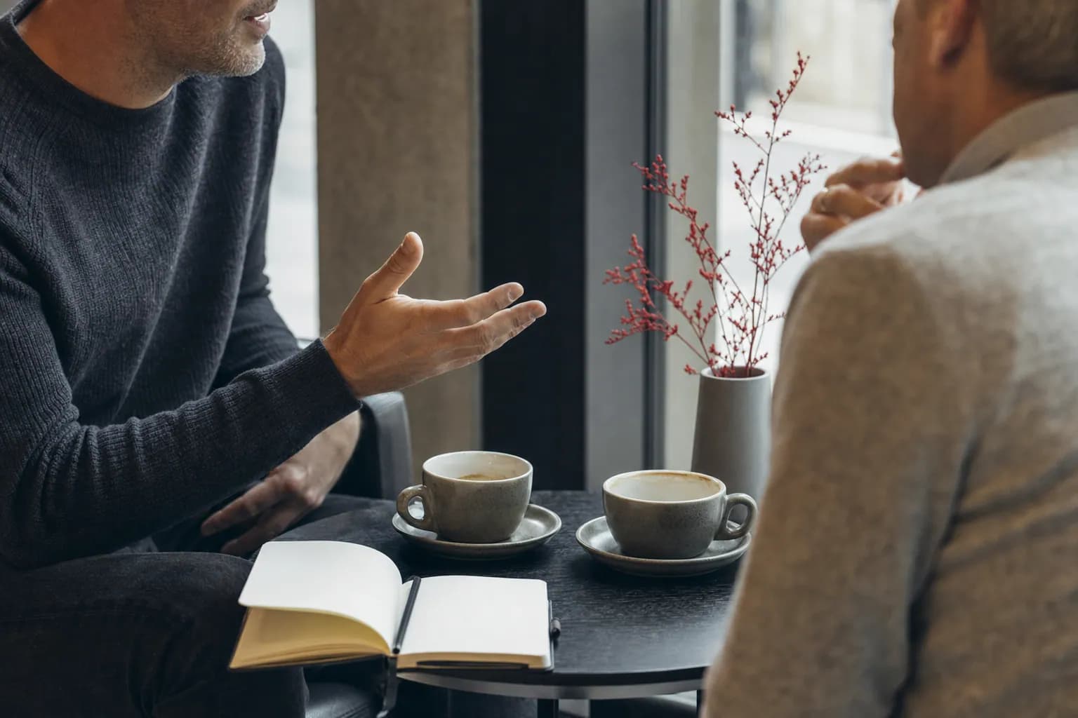 Zwei Kaffeetassen und ein offenes Notizbuch auf einem Cafétisch am Fenster.
