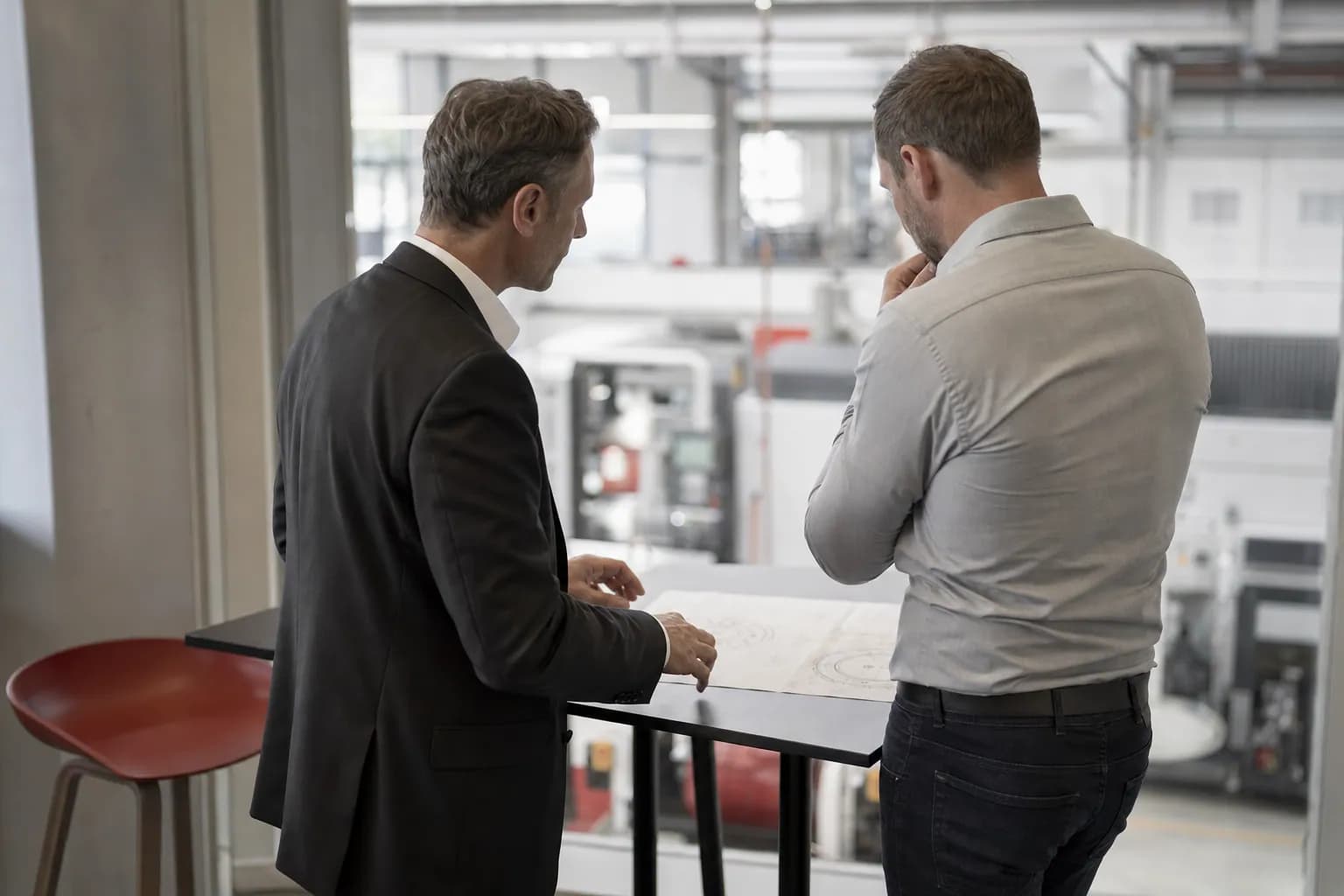 Zwei Männer an einem Stehtisch am Rand einer Maschinenbauhalle prüfen eine technische Zeichnung.